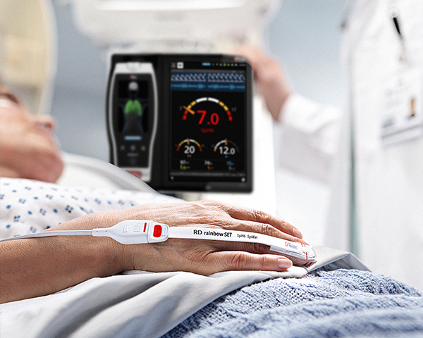 Paciente en una cama de hospital monitorizado con un sensor Masimo con un dispositivo Root y un médico al fondo comprobando un dispositivo Root.
