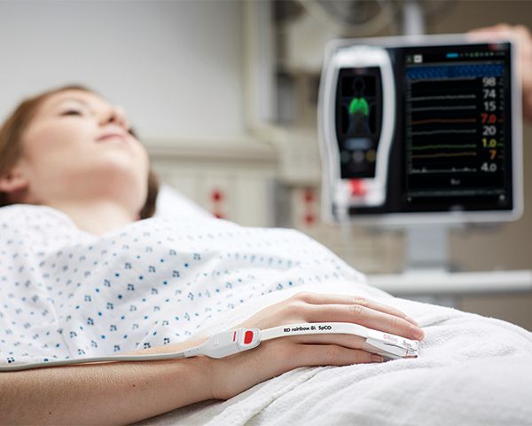 Paciente dormido en la cama del hospital usando un sensor de dedo Masimo con un dispositivo Root al fondo.