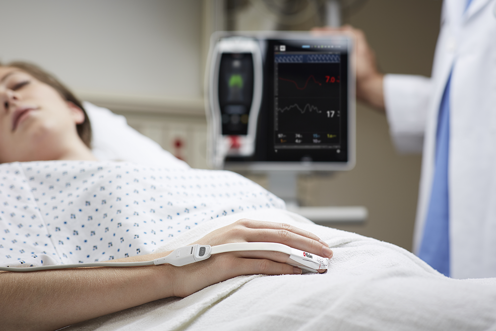 Paciente dormido en la cama del hospital usando un monitor Masimo colocado en el dedo y un dispositivo Masimo Root al fondo.